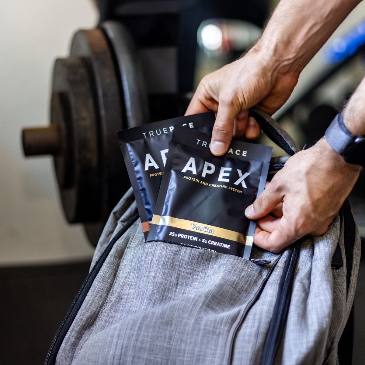A close-up of a person's hands placing two black truePACE APEX protein powder packets into a gray gym bag, with a weight plate visible in the background.