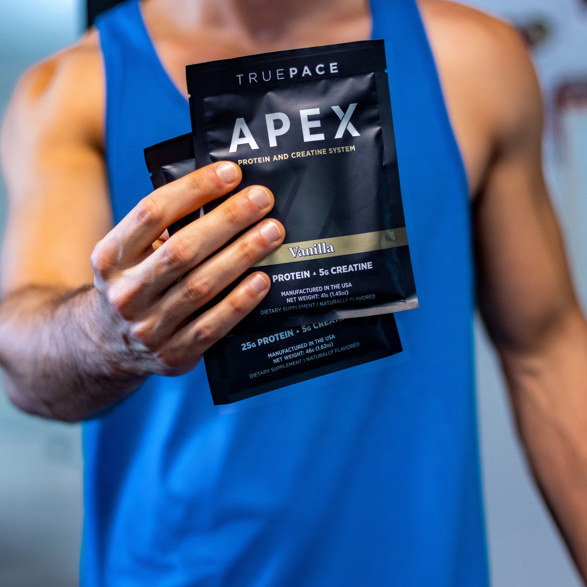 A man in a blue tank top holds two truePACE APEX protein powder packets, showcasing the Vanilla flavor.